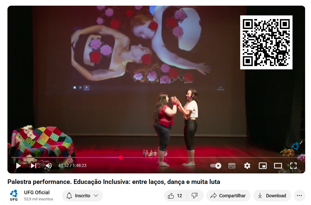 Print de vídeo com palco com luz vermelha. À
esquerda, há um sofá coberto com colcha de retalhos de crochê; ao
centro, duas mulheres em pé, uma de frente para a outra e de mãos dadas.
Ao fundo, uma tela projeta a imagem das duas mulheres deitadas em
posição contrária, com cabeça para o centro. Elas se olham seriamente,
com os braços ao lado do corpo. Usam top preto e, sobre seus cabelos,
rosto, peito, bem como ao seu redor, há rosas vermelhas e rosas de
crochê. No canto superior direito, aparece um QR Code.