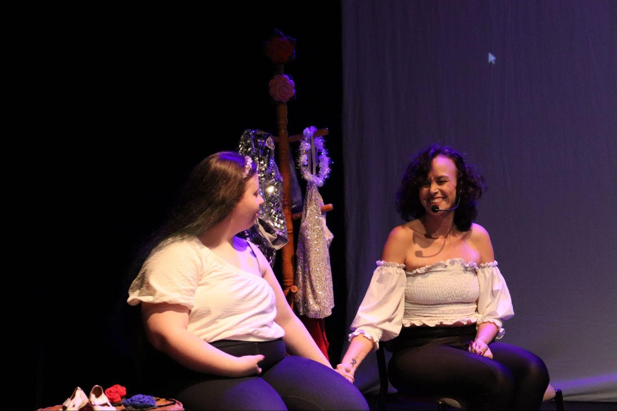 Ao fundo, um cabide com roupas e tiaras. À frente, duas mulheres sorrindo e de mãos dadas. À esquerda, uma mulher com cabelos longos de pontas azuis, pele muito branca, usando blusa branca de um ombro só e calça escura. À direita, uma mulher de cabelo encaracolado na altura dos ombros, usando blusa branca tomara que caia com mangas longas e calça preta. À esquerda, uma pequena mesa com sapatinhos de bebê e rosas de crochê