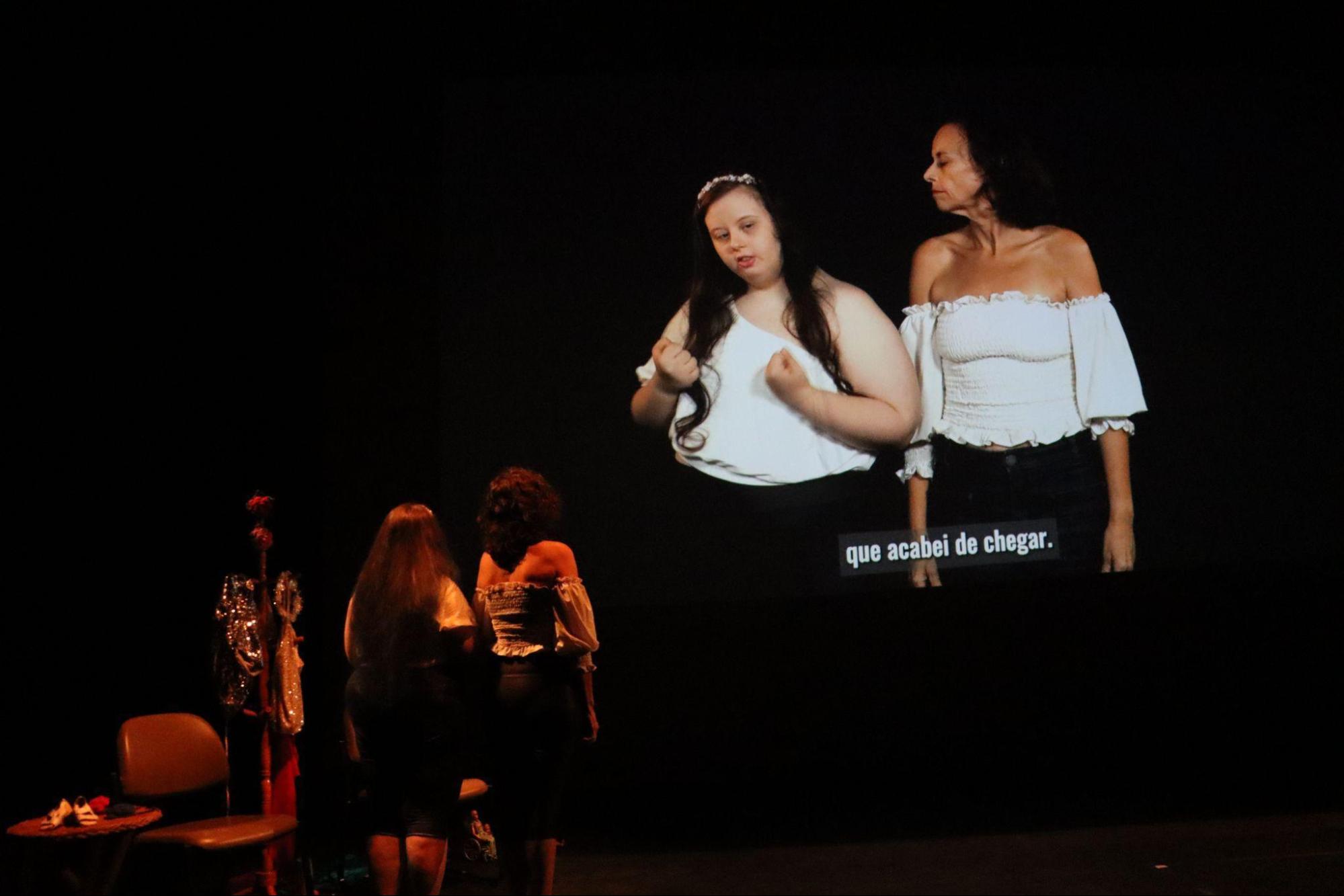 No palco, duas mulheres estão em pé, de costas,
de mãos dadas. Na tela, são projetadas as mesmas mulheres com a mesma
roupa que estão no palco. À esquerda, uma mulher branca com cabelos
longos de pontas azuis, usando blusa branca de um ombro só e calça
escura, gesticula e flexiona os braços. À direita, ao lado, uma mulher
de cabelo encaracolado na altura dos ombros, vestindo blusa branca
tomara que caia com mangas longas e calça preta.
