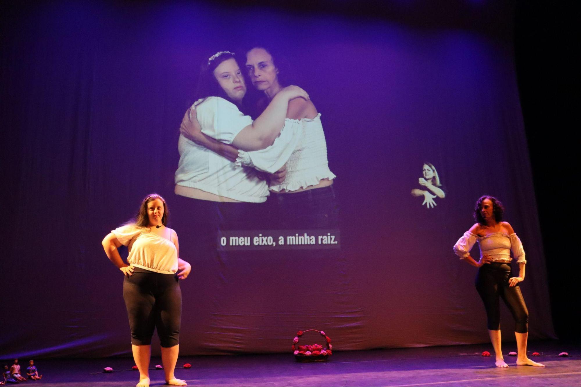 Ao fundo, telão com duas mulheres abraçadas, com
a legenda “o meu eixo, a minha raiz”. À direita, há um intérprete de
Libras. No palco, à frente, duas mulheres posam com uma das mãos na
cintura; elas vestem blusas brancas e calças escuras. No centro do
palco, há uma cesta com flores de crochê.
