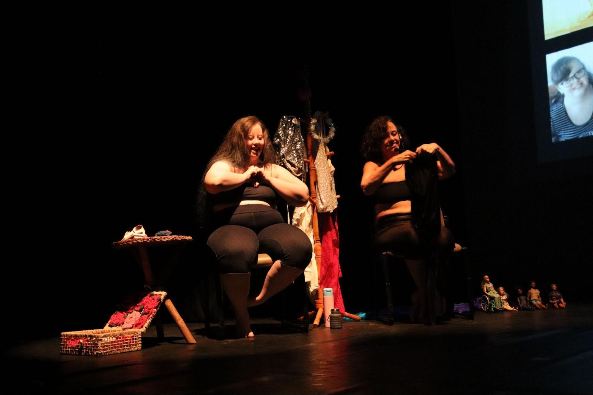 Duas mulheres estão sentadas em cadeiras,
vestindo top e calça escuros. Entre elas, há um com roupas e tiaras. A
mulher à esquerda, de cabelos longos, faz sinal de coração com as mãos;
ao seu lado direito, há uma pequena mesa e uma cesta de flores. A mulher
à direita, de cabelos curtos, segura uma roupa; ao seu lado esquerdo,
estão algumas bonecas.