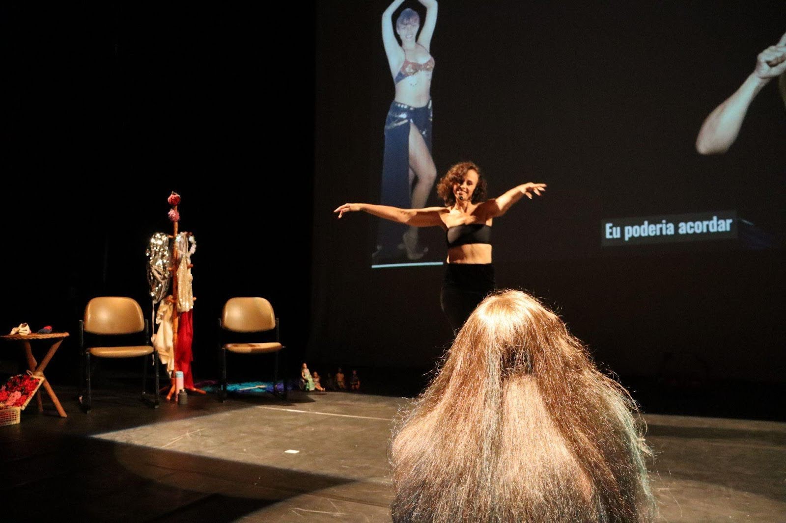 Mulher em pé, com braços abertos, dança diante
de uma tela que exibe imagens ao fundo. À frente, uma jovem está sentada
no chão, olhando a mulher dançar.