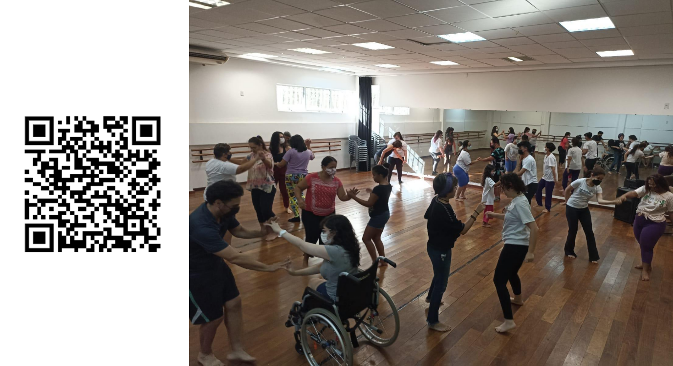 QR code. Sala de aula de dança, à esquerda, uma
barra de madeira; à direita, uma parede com grande espelho. Ao centro,
20 pessoas, com diferentes idades, tamanhos e deficiências, dançam em
duplas.