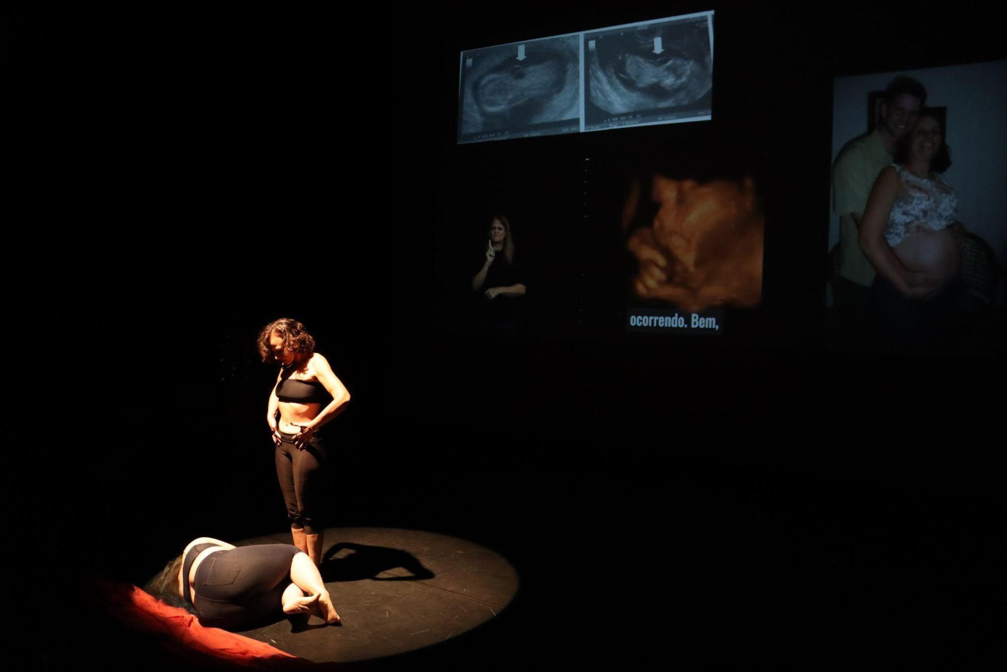 Mulher deitada no chão, encolhida; outra mulher
em pé à sua frente, com a mão sobre o ventre, olhando para baixo. As
duas estão de top e calça escuros.