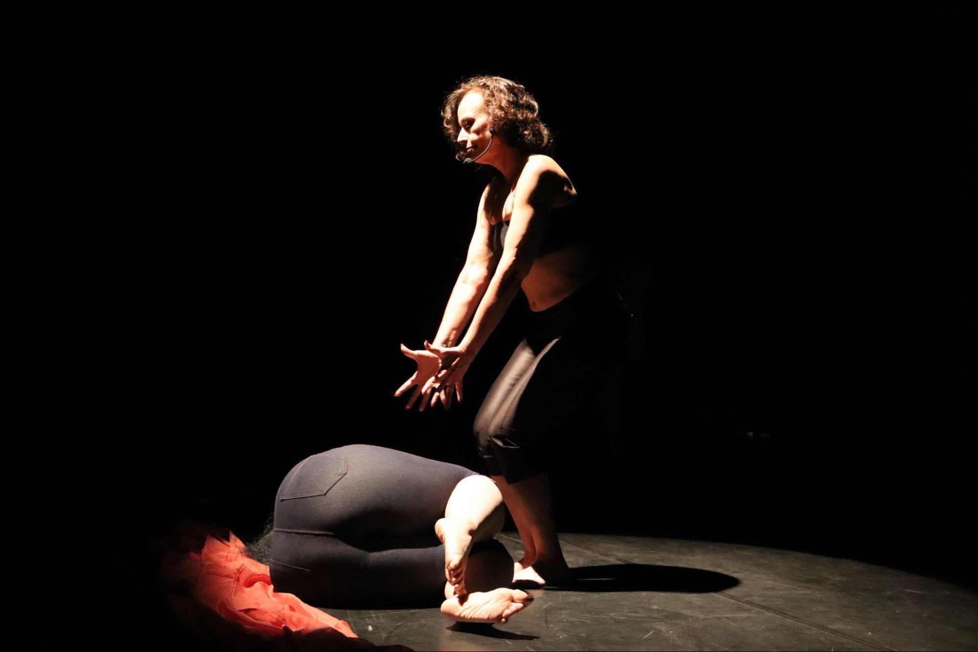 Jovem deitada no chão, encolhida, e outra mulher
em pé à sua frente, com as mãos voltadas para frente e para baixo,
olhando para baixo. Ambas vestem top e calça escura.