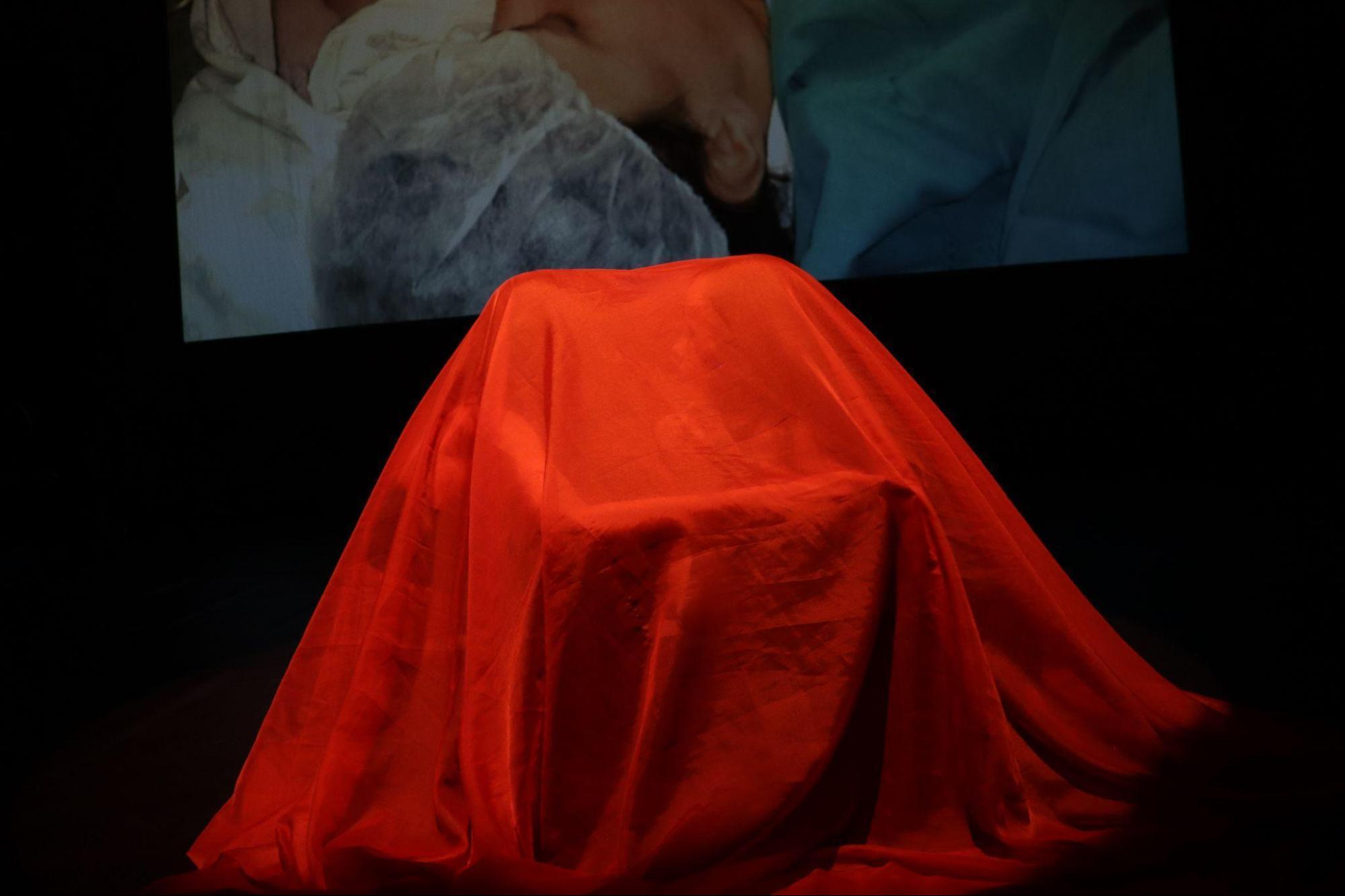 À frente do palco, duas mulheres estão sentadas
no chão, uma de frente para outra, sob um tecido transparente laranja.
Elas se abraçam.