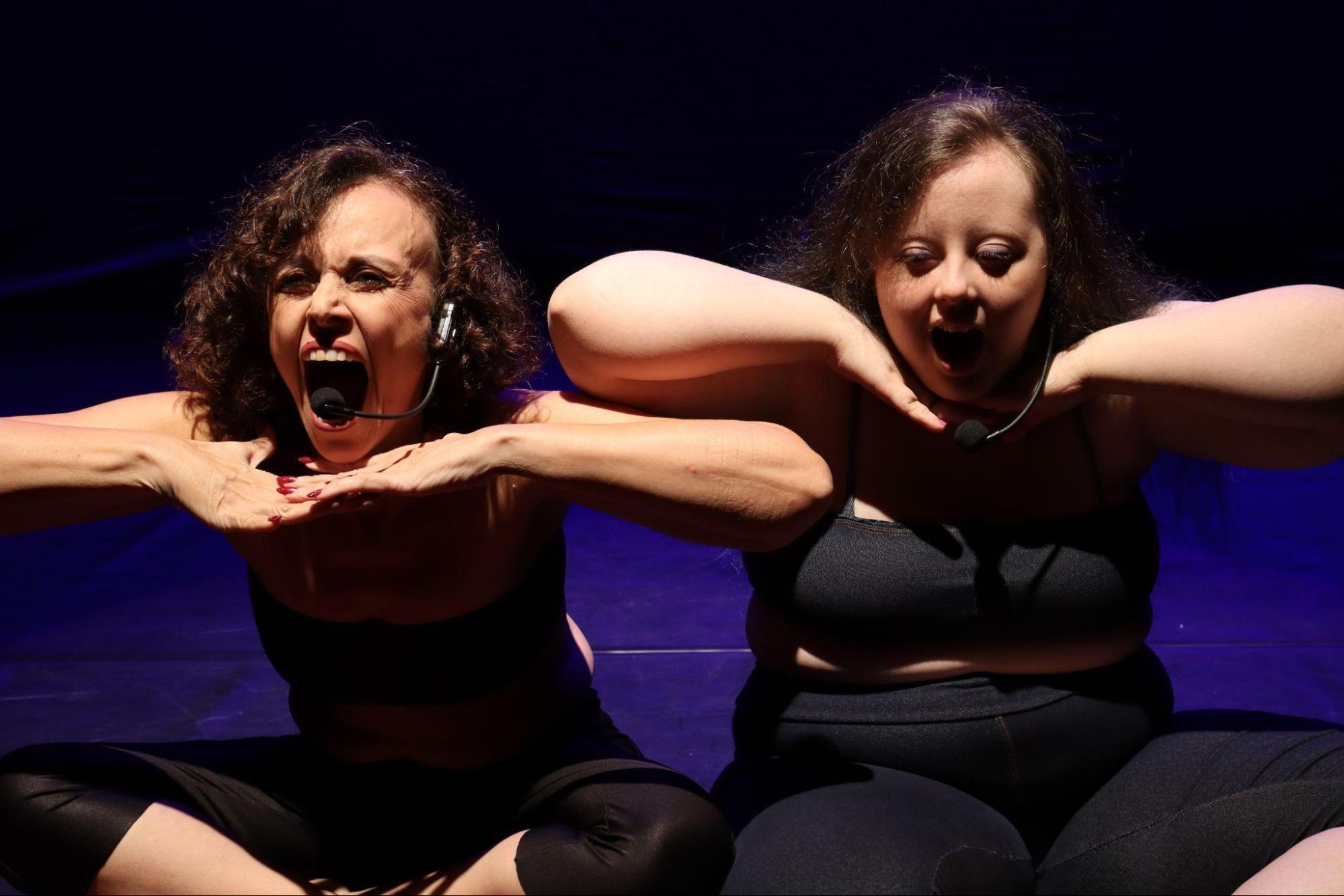 Duas mulheres estão sentadas no chão, com as
mãos apoiadas abaixo do queixo e bocas bem abertas.