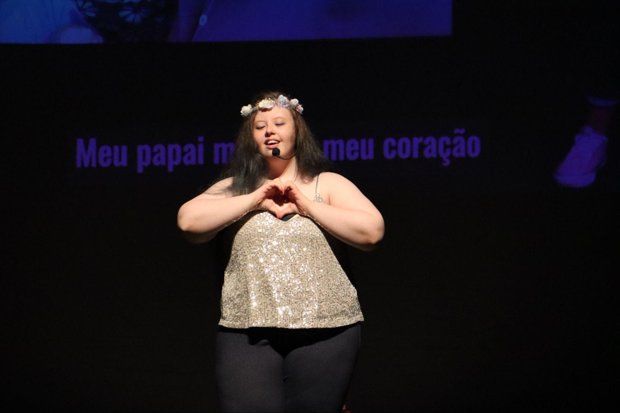 Jovem com síndrome de Down, com coroa de flores
brancas, camisa dourada sem mangas e calça escura, faz um coração com as
mãos. Com um microfone de cabeça, canta. Ao fundo, na tela, aparece a
legenda: “Meu papai mora no meu coração”.