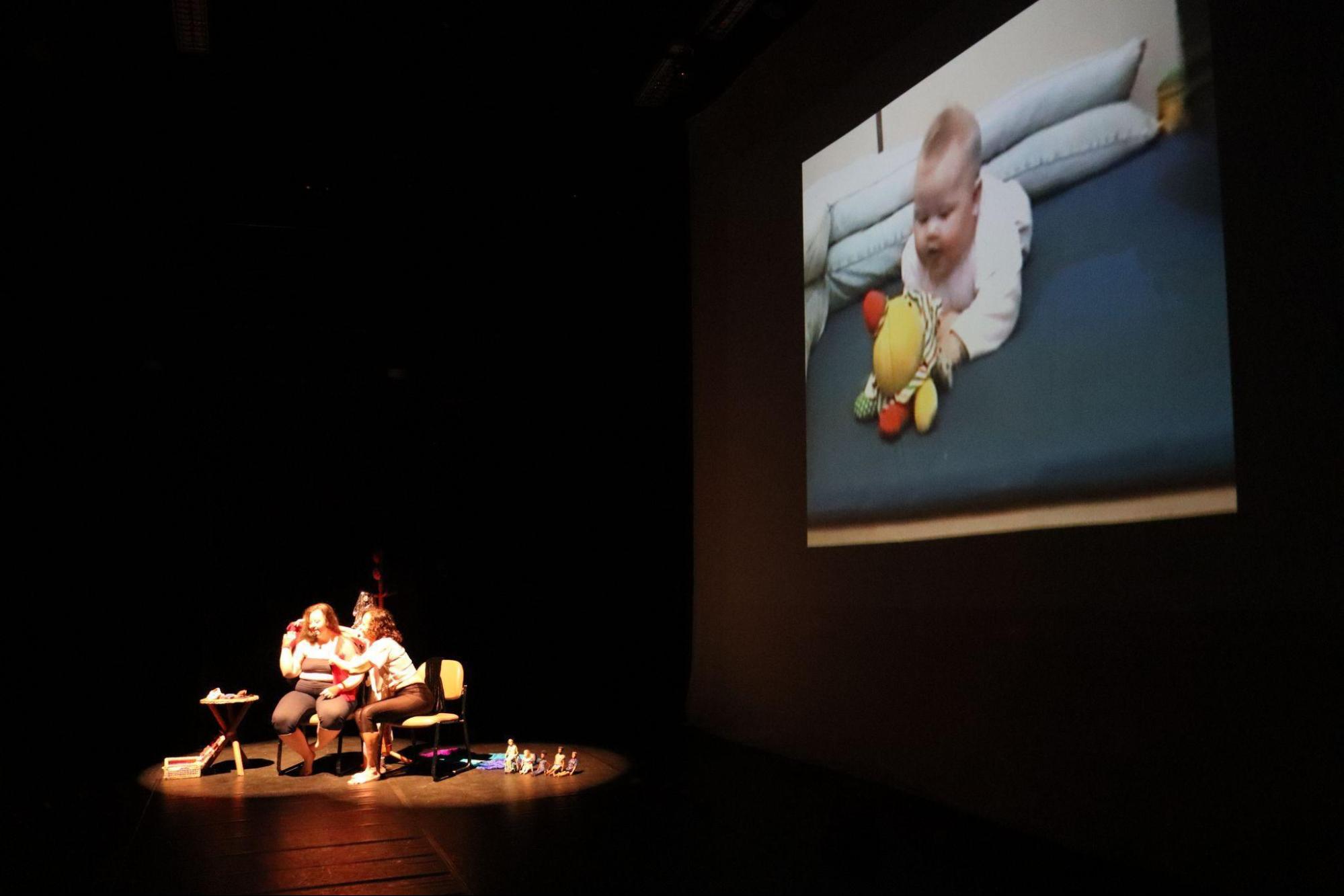 No palco, em foco redondo de luz, duas mulheres
sentadas conversam e colocam blusas. Na tela, atrás, bebê deitada de
barriga para baixo, com brinquedo colorido nas mãos.