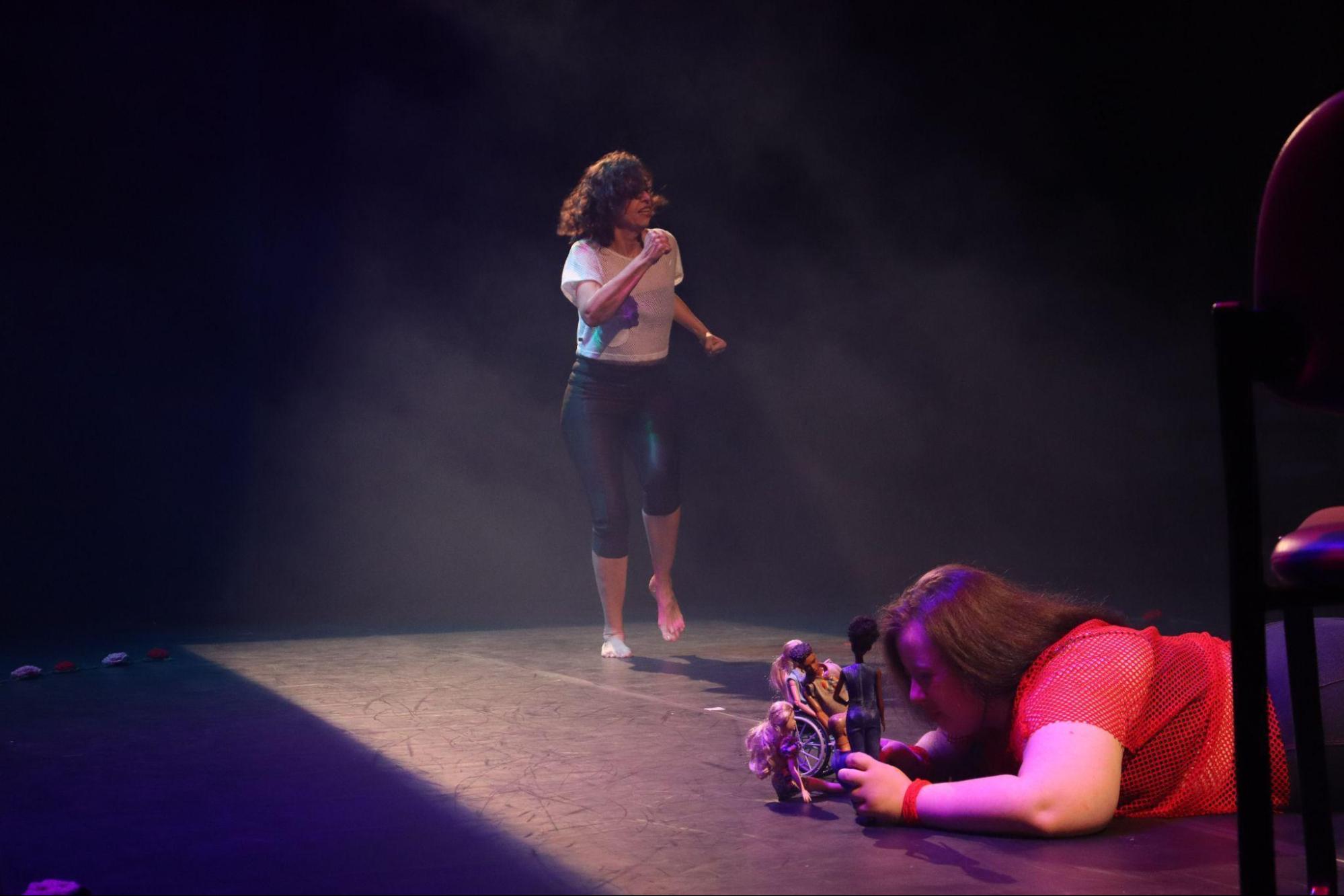 Jovem está deitada no chão, brincando com
bonecas, no canto esquerdo do palco. No centro do palco, a mulher faz
movimento de corrida estacionária. Ambas estão em foco retangular de
luz.