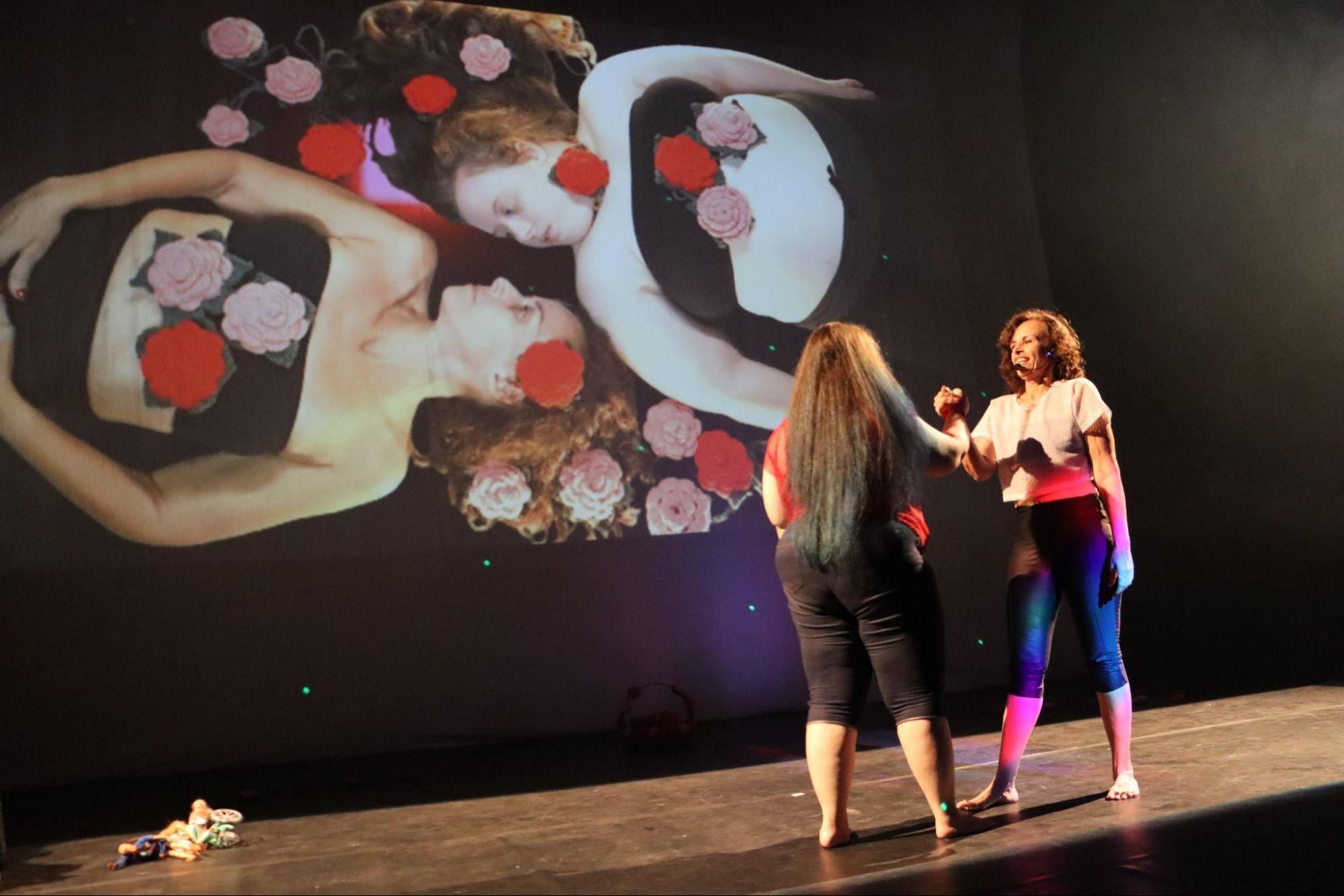 Duas mulheres no centro do palco, em pé, uma de
frente para a outra, de mãos dadas. Atrás, na tela, as mesmas mulheres
deitadas, com rosas de crochê à volta.