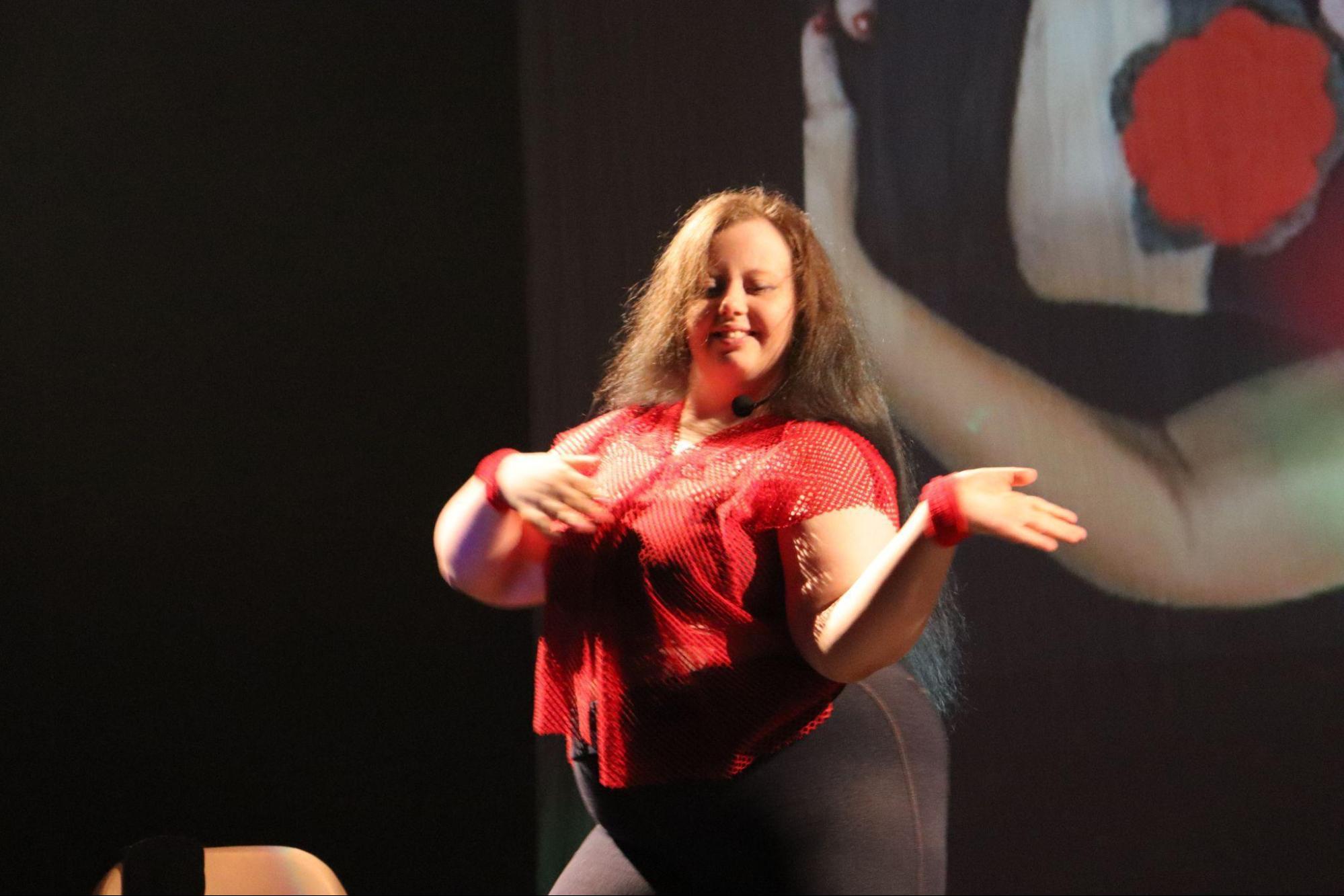 Jovem com síndrome de Down, de cabelos longos,
sorri vestindo blusa vermelha, luvas nos pulsos e calça preta, movimenta
os braços dançando.