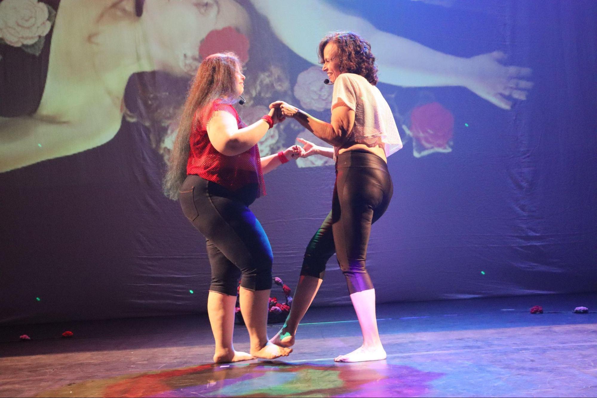 Duas mulheres no centro do palco, em pé, uma de
frente para a outra, de mãos dadas, encostando um pé no pé da outra.