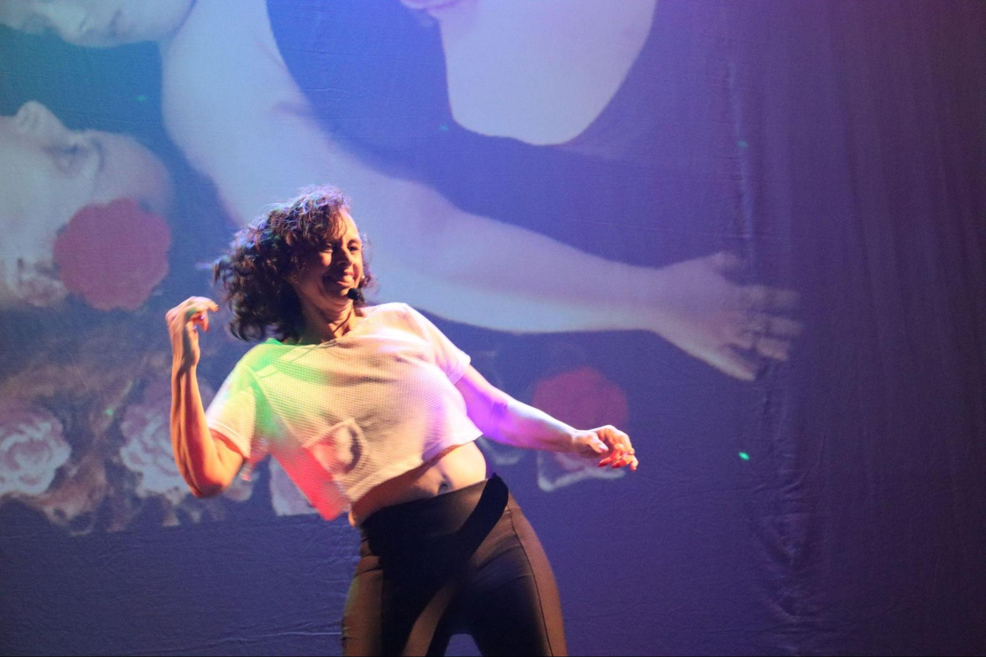 Mulher de cabelos encaracolados curtos, com
camiseta branca curta e calça preta, sorri enquanto dança, movimentando
os braços.