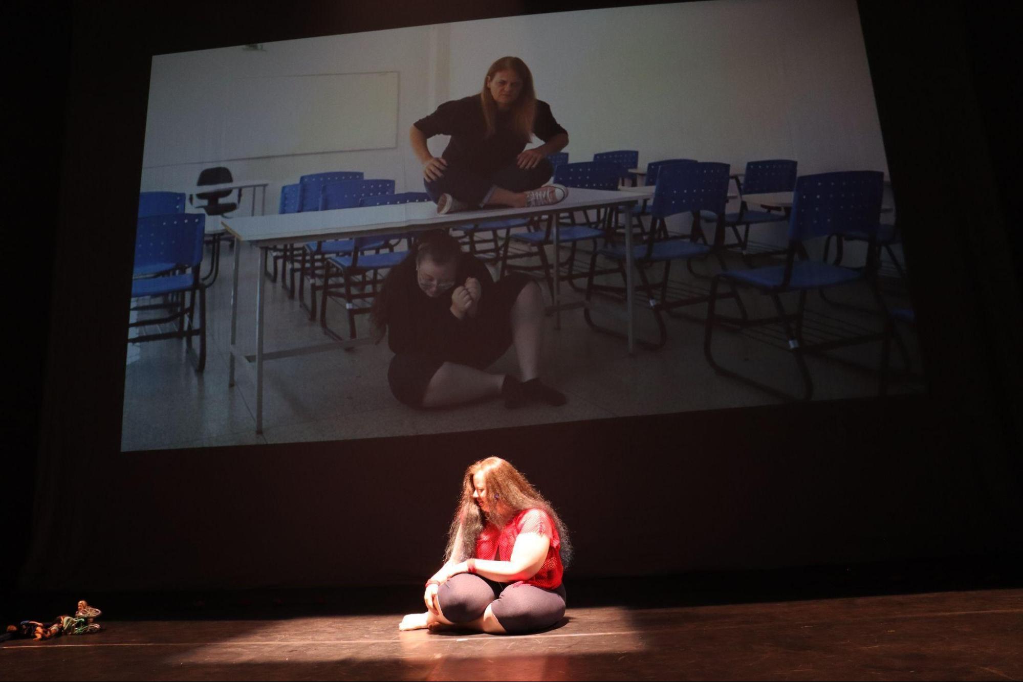 Mulher jovem sentada ao chão, no centro do
palco, em foco de luz, com as pernas flexionadas para o lado esquerdo e
as mãos apoiadas sobre elas, com expressão de sofrimento. Na tela,
atrás, aparece uma sala de aula; embaixo de uma mesa, uma mulher com
cabelo amarrado, blusa de manga comprida, calça e meia pretas; em cima
da mesa, a intérprete de Libras.