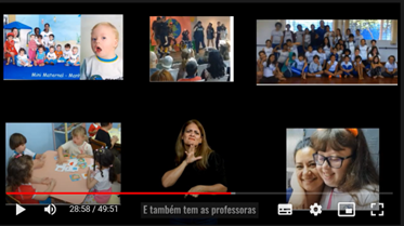 Imagem com fundo preto e cinco fotos, ao centro
intérprete de Libras. A primeira foto, acima à esquerda, mostra uma bebê
muito branca, com cabelo ralo e loiro, olhos azuis e boca aberta,
vestindo camiseta branca com detalhes azuis; ao lado, na mesma foto,
aparecem vários bebês com duas professoras. Abaixo, segunda foto, em uma
mesa quadrada bege, quatro crianças, uma delas, branca, de cabelos
loiros amarrados em um rabo de cavalo distribui peças de jogo para os
demais. Acima, no centro, foto de adolescentes em um palco dançando; um
deles usa cadeira de rodas. Ao lado, à direita, uma turma grande de
adolescentes em foto com a professora. Abaixo, uma adolecente branca, de
cabelo longo e óculos, abraçada com uma mulher que sorri.