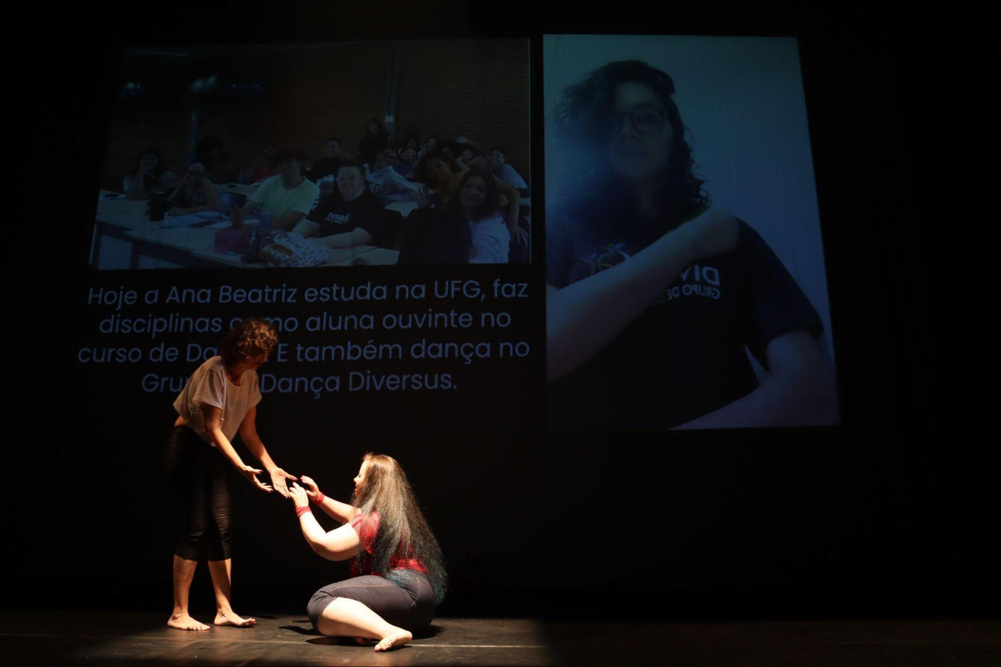 Em foco de luz, à esquerda, mulher em pé, de
camisa branca e calça preta, dá as mãos para a jovem sentada no
chão.