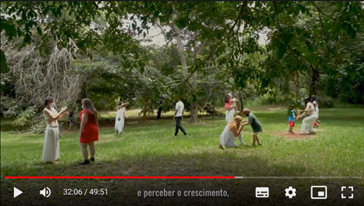 Em um parque com grama verde, ao fundo árvores,
no espaço, mulheres de roupa branca dançam com adolescentes e crianças
de roupas coloridas.