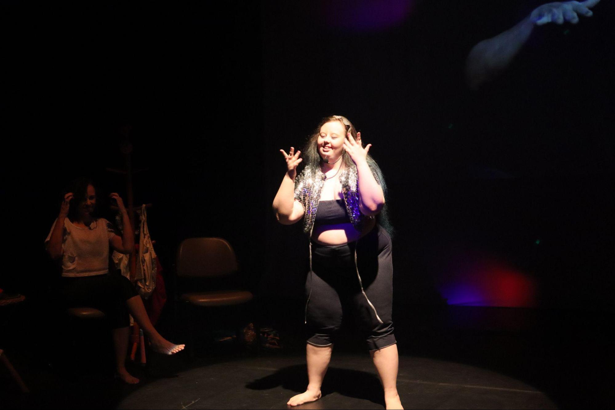 Em foco de luz, jovem com síndrome de Down,
sorrindo, com top e calça preta, blusa prateada brilhante aberta à
frente, com tiara prateada, microfone de cabeça, sorri e movimenta as
mãos. Ao lado esquerdo, uma mulher, na penumbra, a observa.