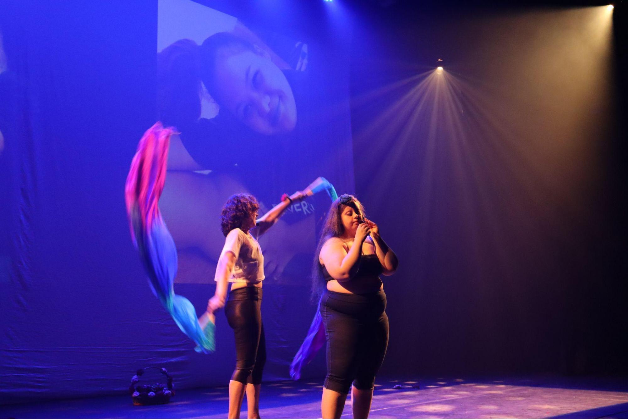 Uma mulher no centro do palco, usando calça e
top escuros, com microfone de lapela, cantando. Atrás dela, outra mulher
ergue os braços, segurando leques com tecidos roxo, rosa e branco que
voam no ar, desenhando um círculo.