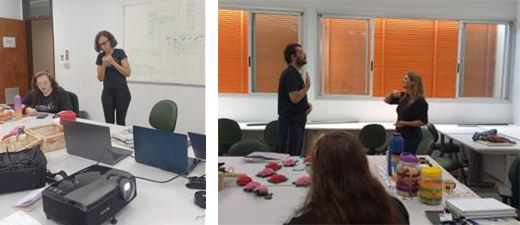 Duas fotos em uma sala de aula com paredes
brancas, porta de madeira, lousa branca toda escrita e janelas com
persianas laranja. Na primeira foto, Vanessa está em pé, vestindo roupa
preta e óculos; Ana Beatriz está sentada à frente de uma mesa branca.
Sobre a mesa, há um datashow ligado, uma cesta de vime com rosas de
crochê, dois computadores, uma garrafa de água e celulares. Na segunda
foto, Ana Beatriz aparece de costas, sentada na mesma mesa, à sua
frente, estão várias flores de crochê cor-de-rosa e vermelhas. Em pé, do
outro lado da mesa, duas pessoas fazem sinais em Libras, uma para a
outra.