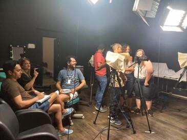 Estúdio de gravação com luzes e paredes pretas.
Há poltronas pretas, três câmeras de vídeo em tripés e uma equipe de
sete pessoas discutindo o vídeo que assistem na câmera. Quatro pessoas
em pé, próximas à câmera, e três sentadas.