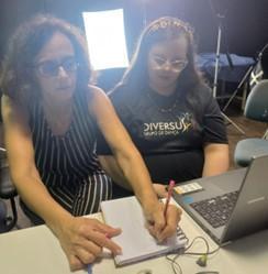 Em um estúdio com paredes pretas revestidas com
isolamento acústico, há uma mesa branca. À frente dela estão sentadas
Ana Beatriz e Vanessa. Vanessa realiza um desenho e escreve
palavras-chave para auxiliar na audiodescrição.