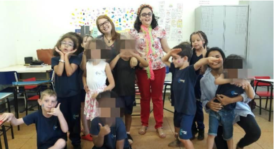 Audiodescrição (AD): da esquerda para direita de cima para baixo temos a imagem da turma reunida, onde a professora e a pesquisadora aparecem mais atrás, algumas crianças agachadas do lado esquerdo, outras crianças e a monitora do lado direito com a criança com TGD apoiada no colo. Ambos aparecem de forma descontraída, alguns sorridentes, outros alegres, mandando beijo, fazendo coração, cada um com uma pose diferente e a seu modo.