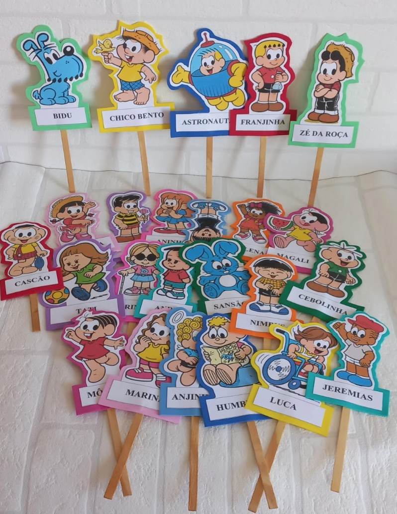 Audiodescrição (AD): imagem contendo vários palitoches com a imagem de personagens do cartunista Maurício de Sousa da Turma da Mônica. Em cada palito, há a imagem de um personagem colado em um fundo colorido recortado contornando a mesma. Neles, há o nome do respectivo personagem: Luca, Jeremias, Cebolinha, Cascão, Sansão, Marina, Magali, Aninha, etc. Alguns estão encostados em uma parede de fundo branco e outros estão dispostos bem juntinhos, uns em cima de parte de outros, na base da mesa com a mesma cor do fundo.