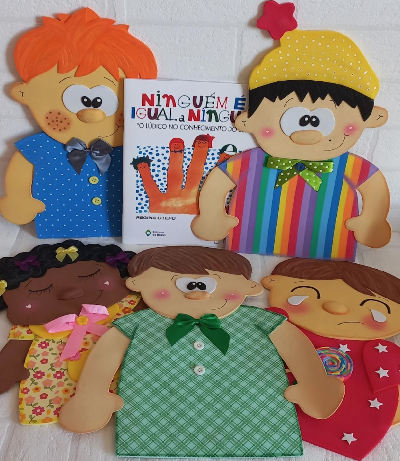 Audiodescrição (AD): imagem contendo Kit com 5 Luvas pedagógicas em E.V.A. com os personagens do livro: Ninguém é igual a ninguém das autoras Regina Otero e Regina Rennó. Encostados na parede de fundo branco estão o livro no centro entre o personagem ruivo de camisa azul com bolinhas brancas à esquerda e o de cabelo preto, usando gorro amarelo de bolinhas brancas, estrela na ponta do gorro vermelha. Veste camisa de listras verticais coloridas. Na base da mesa com a mesma cor do fundo, dispostos bem juntinhos, uns em cima de parte de outros, estão, à esquerda, a menina negra de cabelos enrolados, fitinhas coloridas nos cabelos, olhos fechados, veste vestido amarelo com flores rosa em tons diferentes. O menino de cabelos castanhos, olhos pretos, veste camisa verde com listras finas verdes na diagonal e usa gravata verde escura. Do lado direito, um menino chora e segura um pirulito na mão direita. Veste camiseta vermelha com estrelas brancas.